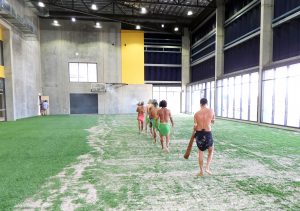 Indigenous group performing a traditional ceremony with a didgeridoo inside the building with astro turf and large windows